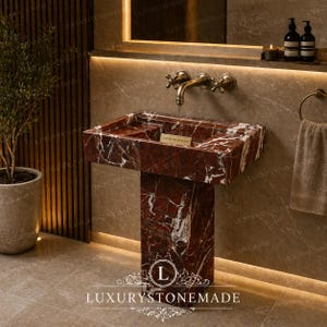Peut inclure: Un lavabo sur pied en marbre rouge veiné de blanc. Le lavabo a un bassin rectangulaire et un socle assorti. Un robinet de style vintage est monté sur le mur au-dessus du lavabo. Le texte "LUXURYSTONEMADE" est visible.
