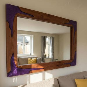 May include: A rectangular wooden mirror with a dark brown frame and purple resin accents in the corners. The mirror reflects a room with a window, curtains, and a sofa. The mirror is mounted on a light-colored wall.
