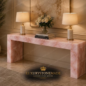May include: A pink marble console table with two matching lamps and a floral arrangement. The lamps have beige shades and gold bases. The table has a rectangular shape and is made of pink marble. The text "LUXURYSTONEMADE" is at the bottom.