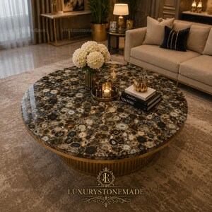 May include: A large, round coffee table with a polished, patterned stone top. The stone features a mix of brown, black, and cream colors. The table has a gold-colored, ribbed base and a gold trim. A vase of white flowers, books, and a candle sit on top.