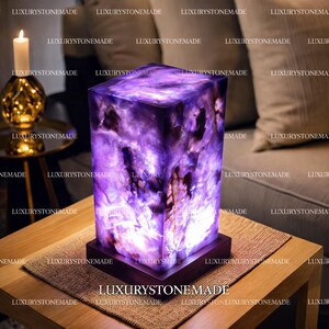 May include: A rectangular purple stone lamp with a dark wooden base. The lamp is lit, revealing the natural patterns within the stone. The lamp sits on a wooden table, with a candle and gold ornament in the background.