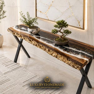Könnte beinhalten: Eine moderne Konsolentisch mit einer Tischplatte aus Holz mit natürlicher Kante, mit einem Design aus schwarzem und weißem Epoxidharz und Goldblattakzenten. Der Tisch steht auf schwarzen Metallbeinen in X-Form. Zwei kleine Topfpflanzen stehen darauf.