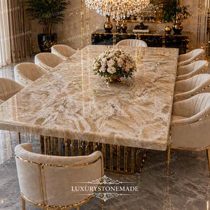 May include: A large, rectangular dining table with a light-colored, patterned stone top. The table is surrounded by ten beige upholstered chairs with gold trim. A floral arrangement sits in the center. The table base is gold.