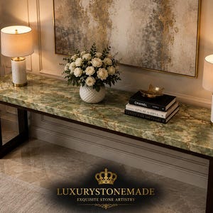 May include: A long, rectangular green marble table with dark brown legs. Two lamps with beige shades and white bases flank the table. A vase of white roses, books, and decorative items are on the table. The text "LUXURYSTONEMADE" is at the bottom.