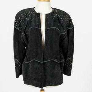 May include: Black suede jacket with silver-toned stud embellishments on the shoulders and cuffs. The jacket has a collarless design and a subtle, wavy seam detail. The jacket is displayed on a mannequin.