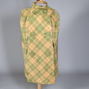 May include: A double-breasted cape featuring a plaid pattern in shades of sage green, cream, and orange. The garment has a high collar, structured shoulders, and six large, round, fabric-covered buttons arranged in two vertical rows down the front.
