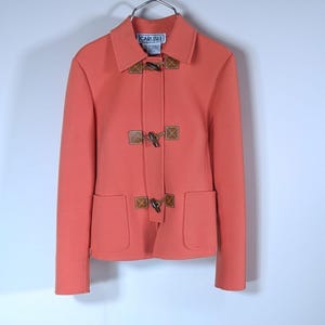 May include: Coral-colored jacket with toggle closures and two front pockets. The jacket has a collared neckline and long sleeves. The brand name "CARLISLE" is visible on a label at the neckline. The jacket is hanging on a metal hanger.