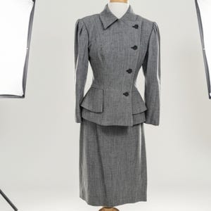 May include: A vintage gray tweed suit displayed on a mannequin. The suit features a fitted jacket with a peplum, a notched collar, and black buttons. The matching skirt is knee-length. The suit is a classic example of mid-century fashion.