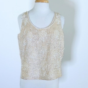 May include: A sleeveless, cream-colored top fully embellished with beads and sequins. The scoop-neck top is displayed on a mannequin. The intricate beadwork creates a textured surface with a subtle, shimmering effect. The design is reminiscent of vintage or formal wear.