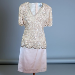 May include: A vintage, knee-length dress in a soft pink and beige color scheme. The bodice features a V-neck and is adorned with sequined floral patterns. The skirt is a solid, lighter pink, creating a two-tone effect. The dress has short sleeves.