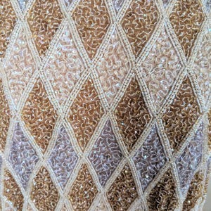 Puede incluir: Primer plano de una tela brillante y texturizada con un patr&oacute;n de diamantes. El dise&ntilde;o presenta secciones alternas de lentejuelas y cuentas doradas, plateadas y crema, creando una apariencia lujosa y ornamentada. La tela probablemente se usa para ropa formal.