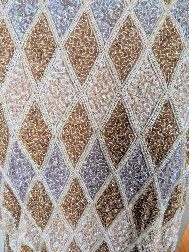 Puede incluir: Primer plano de una tela brillante y texturizada con un patr&oacute;n de diamantes. El dise&ntilde;o presenta secciones alternas de lentejuelas y cuentas doradas, plateadas y crema, creando una apariencia lujosa y ornamentada. La tela probablemente se usa para ropa formal.