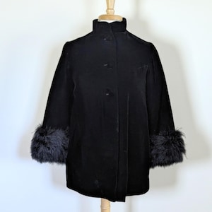 May include: A black velvet coat with a high collar and three buttons down the front. The sleeves are three-quarter length and finished with black feather cuffs. The coat is displayed on a wooden dress form against a white background.