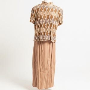 Puede incluir: Un conjunto vintage de dos piezas con una blusa de manga corta y una falda larga. La blusa tiene un cuello alto y un estampado de rombos en tonos marr&oacute;n, beige y dorado. La falda es de color marr&oacute;n claro liso y fluida.