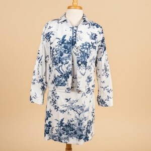 May include: A two-piece set consisting of a long-sleeve button-up jacket and a matching skirt, both featuring a white base with a detailed blue toile de jouy pattern depicting pastoral scenes, foliage, and birds throughout the entire garment.