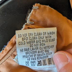 Puede incluir: Primer plano de una etiqueta de prenda con instrucciones de cuidado en ingl&eacute;s y franc&eacute;s. La etiqueta es blanca con texto negro, sujeta a una tela marr&oacute;n claro. Se ve una cremallera.
