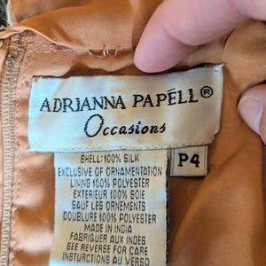 Puede incluir: Primer plano de una etiqueta de una prenda Adrianna Papell Occasions. La etiqueta es blanca con texto negro, incluyendo el nombre de la marca, "Occasions", y detalles de la composici&oacute;n de la tela. La talla es P4. La prenda es de color melocot&oacute;n.