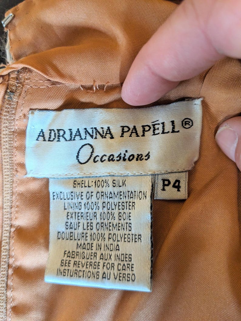 Puede incluir: Primer plano de una etiqueta de una prenda Adrianna Papell Occasions. La etiqueta es blanca con texto negro, incluyendo el nombre de la marca, "Occasions", y detalles de la composici&oacute;n de la tela. La talla es P4. La prenda es de color melocot&oacute;n.