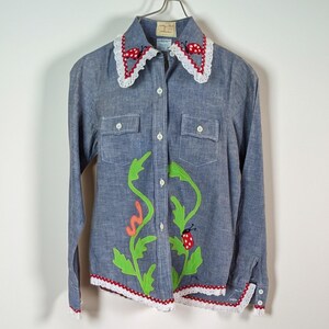 May include: Blue denim shirt with long sleeves, two chest pockets, and a white crochet collar with red polka dots. The shirt features a green and orange applique design with a ladybug.