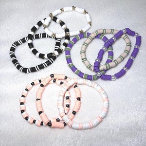 May include: A collection of colorful beaded bracelets. The bracelets feature a variety of colors, including black and white, purple and green, and pink and white. Some bracelets have gold-colored beads or faux pearls.