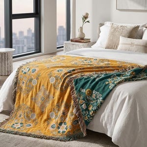 Puede incluir: Una manta mostaza y verde azulado con un estampado floral está sobre una cama. La manta tiene un borde con flecos y está hecha de un material suave y tejido. La cama está cubierta con sábanas blancas.