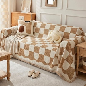 May include: A sofa with a brown and cream checkered throw blanket and decorative pillows. The throw has a scalloped edge with fringe. A cream-colored knot pillow and a decorative pillow with abstract shapes are on the sofa.