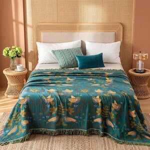 Puede incluir: Una manta turquesa con un estampado de pájaros y flores está sobre una cama. La manta tiene un borde con flecos y está decorada con pájaros y flores amarillas, marrones y azules. Hay almohadas blancas y una almohada verde en la cama.