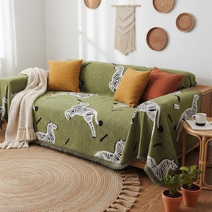 May include: A green sofa cover with a zebra pattern, paired with mustard and rust-colored throw pillows. A cream-colored blanket with fringe is draped over the arm. The room features a jute rug and decorative wall hangings.