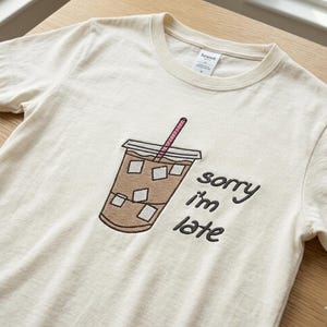 Puede incluir: Camiseta color crema con un diseño bordado de café helado y una pajita rosa. El texto "sorry I'm late" está impreso en negro junto a la taza de café. La camiseta tiene cuello redondo y mangas cortas.
