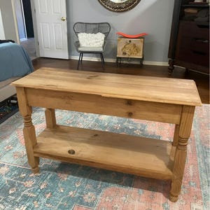 Puede incluir: Una consola de madera marrón claro con un estante inferior y patas torneadas. La mesa está hecha de madera sin terminar y se encuentra sobre una alfombra estampada con colores azul, rosa y crema. La mesa tiene un cajón.