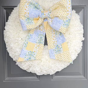 May include: A white, ruffled wreath with a large bow. The bow is made of fabric with yellow floral and blue floral patterns. The wreath is hanging on a grey door.