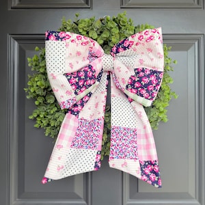 May include: A decorative bow made of patchwork fabric, featuring pink floral, polka dot, and gingham patterns. The bow is attached to a green wreath, creating a charming door decoration. The bow's design includes a mix of textures and colors.