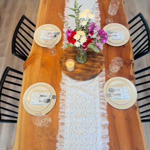May include: A wooden dining table set for a meal. The table is adorned with a white and blue lace runner, plates, silverware, and wine glasses. A vase of flowers and a lit candle are in the center. Black chairs surround the table.