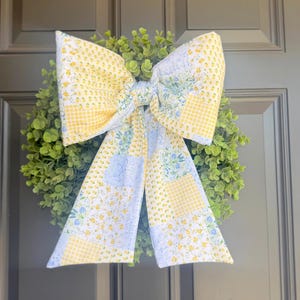 Puede incluir: Una corona decorativa con un gran lazo de tela. El lazo presenta un diseño de patchwork con patrones florales amarillos, blancos y azules. La corona está compuesta de follaje verde, ideal para la decoración del hogar.