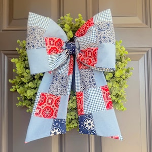 May include: A decorative bow with a patchwork design in red, white, and blue, attached to a green wreath. The bow features a gingham pattern and paisley-style prints. This festive accent is suitable for a front door or interior decor.