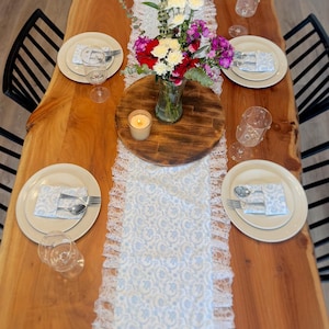 May include: A wooden dining table set for a meal. The table is adorned with plates, silverware, and wine glasses. A floral centerpiece and a lit candle sit on a wooden round board. A blue and white lace table runner adds a decorative touch.