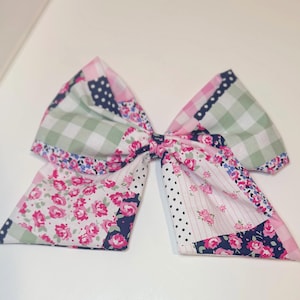以下が含まれることがあります： 様々な柄が組み合わされた、大きなパッチワーク生地のリボン。ピンク、白、緑、ネイビーブルーの生地を使用。ギンガムチェック、花柄、水玉模様、無地など。ヘアアクセサリーや装飾品として使用される可能性が高いリボンです。