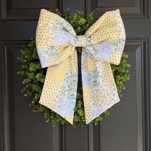 Puede incluir: Una corona decorativa con un gran lazo de tela estampada amarilla y azul. El lazo está centrado en una corona verde y frondosa, creando una decoración de puerta acogedora. La puerta está pintada de gris oscuro.