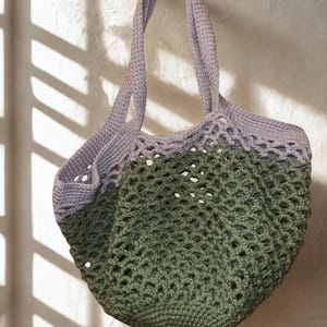 May include: A handmade crochet tote bag in olive green and lavender. The bag features a textured, open weave design and sturdy handles. The bag is hanging against a white wall, with shadows from a window.