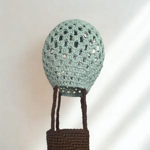 May include: A handmade crochet hot air balloon in a soft teal color. The balloon's basket is a dark brown, and the balloon has an open, airy design. The crochet work is detailed, creating a unique decorative piece.