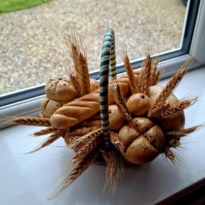 Op de afbeelding: Een mand gevuld met verschillende soorten brood, waaronder broodjes en broden, en tarwearen. De mand heeft een groen-wit gestreepte handgreep. Het brood is lichtbruin van kleur.