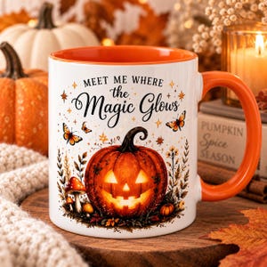 May include: A white ceramic mug with an orange interior and handle, featuring a central glowing jack-o'-lantern, mushrooms, and butterflies. The text reads, "MEET ME WHERE the Magic Glows," surrounded by small star and leaf illustrations.