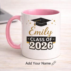 Puede incluir: Taza de cerámica blanca con interior y asa rosas. La taza presenta un gráfico de birrete, el nombre "Emily" en letras doradas y "CLASS OF 2026" en negro. El texto "Add Your Name" está en la parte inferior.