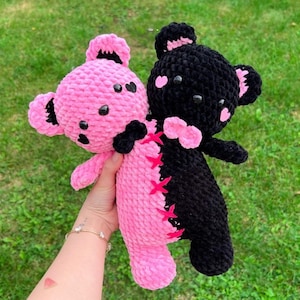 May include: Two handmade crochet teddy bears. One is pink with black accents and the other is black with pink accents. The bears are hugging each other. The pink bear has black heart-shaped eyes and the black bear has pink heart-shaped accents.