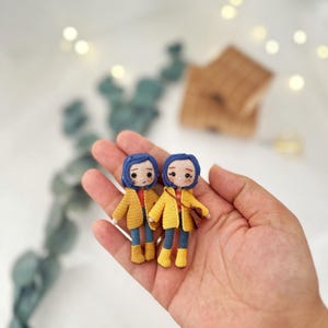 Könnte beinhalten: Zwei handgefertigte Häkelpuppen mit blauen Haaren, gelben Jacken, roten Hemden, blauen Hosen und gelben Stiefeln. Die Puppen werden in einer Hand gehalten, mit einem verschwommenen Hintergrund.