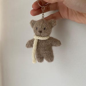 May include: A small, knitted teddy bear keychain in shades of brown and beige. The bear has a cream-colored scarf and a silver keyring. The bear is approximately 3 inches tall.