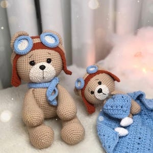 May include: Two crocheted teddy bears. One is upright, wearing a brown aviator hat with blue goggles and a blue scarf. The other bear is lying down, wearing a matching hat and holding a blue blanket. The bears are tan with black eyes.