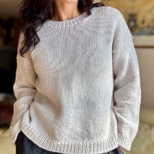 May include: A light grey knitted sweater with a crew neck and long sleeves. The sweater features a ribbed hem and cuffs. The knitwear appears to be a soft, comfortable material. The sweater is a neutral colour, suitable for various outfits.