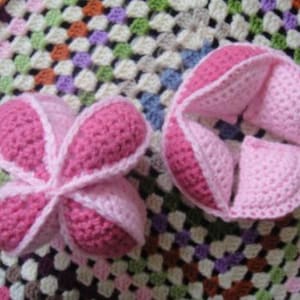 Puede incluir: Dos juguetes de bebé hechos a mano con ganchillo en tonos de rosa. Uno es una bola cerrada y el otro es una bola abierta. Los juguetes están hechos con una puntada texturizada y tienen un borde rosa claro. El fondo es una colorida manta de ganchillo.