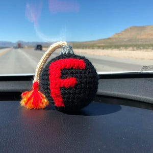 以下が含まれることがあります： 黒い手編みの爆弾型オーナメント。前面に大きな赤い「F」があります。銀色のトップ、ベージュの紐、赤とオレンジのタッセルが付いています。オーナメントは、背景に高速道路がある暗いダッシュボードの上にあります。